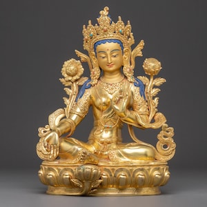 May include: A golden statue of a seated deity with a serene expression. The figure wears an ornate crown and jewelry, with blue accents on the shoulders. The statue is adorned with lotus flowers and intricate details, showcasing a sense of peace and spirituality.