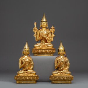 Puede incluir: Tres estatuas budistas doradas. La estatua central está sentada y flanqueada por dos estatuas más pequeñas. Cada figura lleva un sombrero puntiagudo y túnicas ornamentadas. Las estatuas están sobre bases decorativas, sobre un fondo gris.