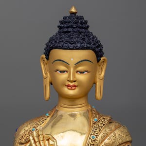 K&ouml;nnte beinhalten: Eine goldene Buddha-Statue mit einem detaillierten, verzierten Gewand und einem ruhigen Ausdruck. Die Statue ist aus Metall und hat ein traditionelles Design.