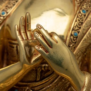 K&ouml;nnte beinhalten: Nahaufnahme der H&auml;nde einer goldenen Statue in Gebetsposition. Die H&auml;nde sind mit aufwendigen Schnitzereien verziert und mit t&uuml;rkisfarbenen Edelsteinen besetzt.