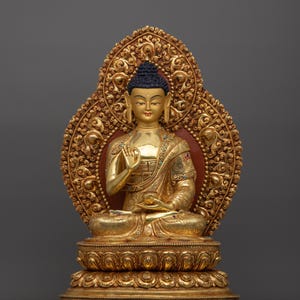 K&ouml;nnte beinhalten: Eine goldfarbene Buddha-Statue, die in meditativer Haltung sitzt. Die Statue ist mit aufwendigen Details verziert und vor einem grauen Hintergrund platziert.