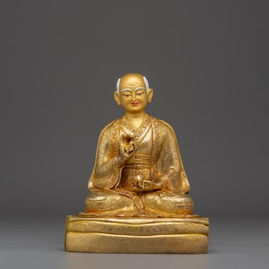 May include: A golden statue of a seated figure with a serene expression. The figure is wearing a detailed robe and is seated on a rectangular base. The statue has a bald head with white eyebrows and a serene expression.