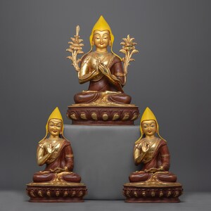 Puede incluir: Tres estatuas budistas tibetanas doradas y rojas con sombreros amarillos. Las estatuas están sentadas sobre una base roja y dorada.