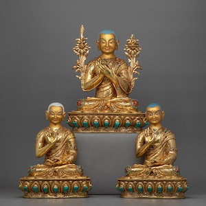 Puede incluir: Tres estatuas doradas de figuras budistas sentadas en una postura meditativa. Las estatuas están adornadas con piedras preciosas turquesa y rojas. La estatua central es más grande que las dos estatuas laterales.