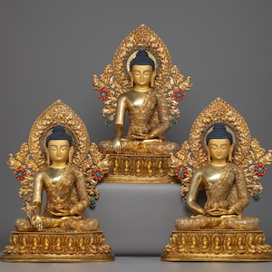 May include: Three gold Buddha statues in a seated meditation pose. Each statue has a blue hair bun and is decorated with detailed gold patterns. The statues are set against a grey background.