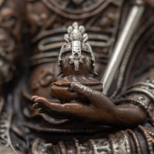 May include: Close-up of a bronze statue's hand holding a small, ornate, silver-colored figure with a crown. The statue is detailed with intricate carvings and a silver-colored trim.