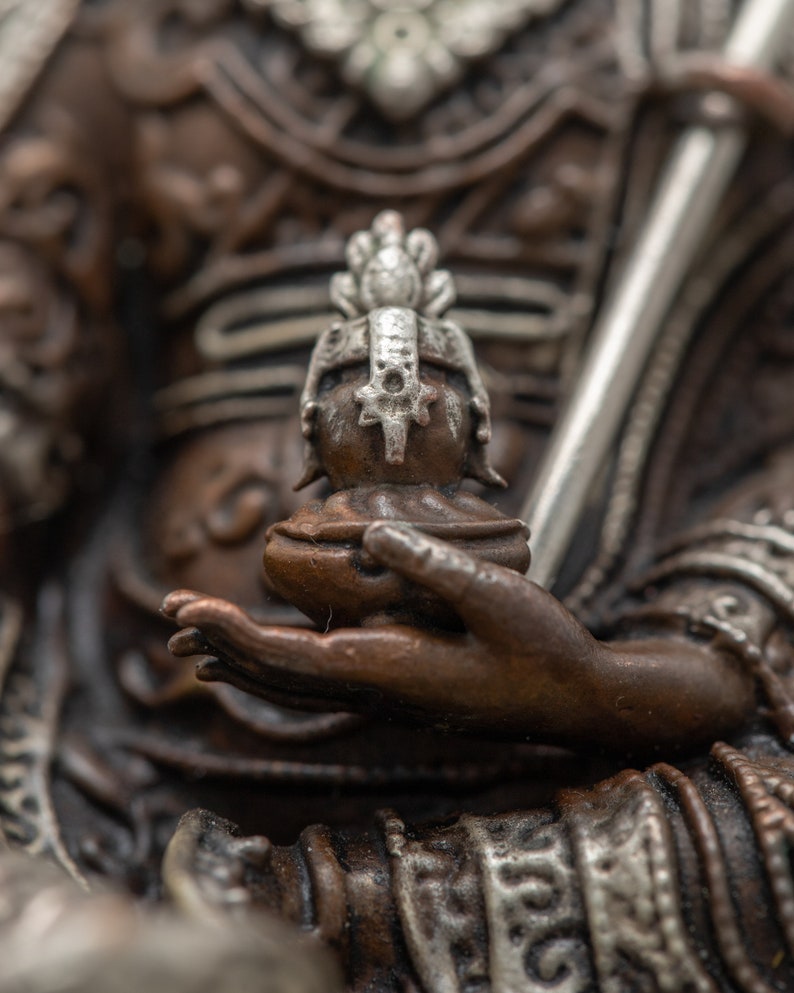 May include: Close-up of a bronze statue's hand holding a small, ornate, silver-colored figure with a crown. The statue is detailed with intricate carvings and a silver-colored trim.