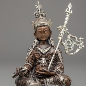 May include: A bronze and silver statue of a deity sitting in a meditative pose. The deity is wearing a crown and ornate clothing. The statue is holding a scepter in one hand and a small object in the other.