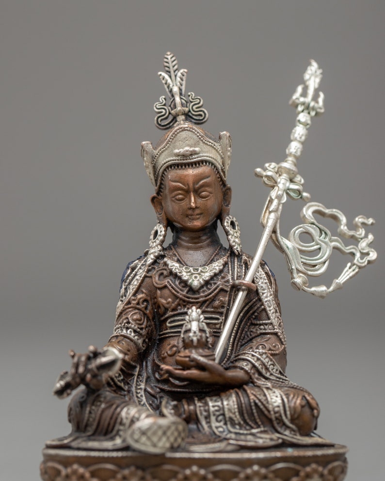 May include: A bronze and silver statue of a deity sitting in a meditative pose. The deity is wearing a crown and ornate clothing. The statue is holding a scepter in one hand and a small object in the other.