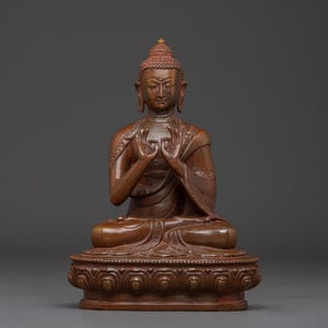 May include: A seated brown Buddha statue with hands in a meditative gesture. The statue is on a decorative base and has a serene expression. The statue is made of a dark brown material.