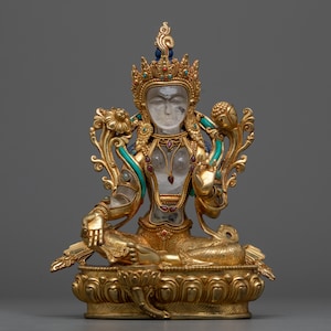 May include: A gold-colored statue of a deity, possibly a Buddhist deity, sitting in a meditative pose. The statue is made of a white stone and has intricate details, including a crown, jewelry, and a lotus base. The statue is adorned with turquoise and red gemstones.