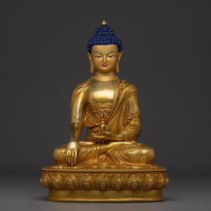 May include: A gold Buddha statue, seated with a blue-coloured hair and a calm expression. The statue features detailed carvings and rests on an ornate base. This statue is a representation of Buddhist art and culture.
