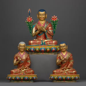 May include: Three gold-painted statues of Buddhist figures in a seated meditation pose. The figures are wearing red and gold robes and are adorned with intricate details. The statues are set against a grey background.