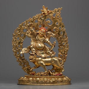 May include: A gold-colored statue of a deity riding a tiger, with a decorative flame-like halo behind them. The statue is detailed with red and blue paint accents.