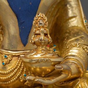 K&ouml;nnte beinhalten: Nahaufnahme einer goldenen religi&ouml;sen Statue, die eine kleine, verzierte Schale h&auml;lt. Die Statue weist komplizierte Details auf, darunter ein flammenartiges Design auf der Schale und t&uuml;rkisfarbene Akzente. Der Hintergrund ist ein weiches, verschwommenes Gold.