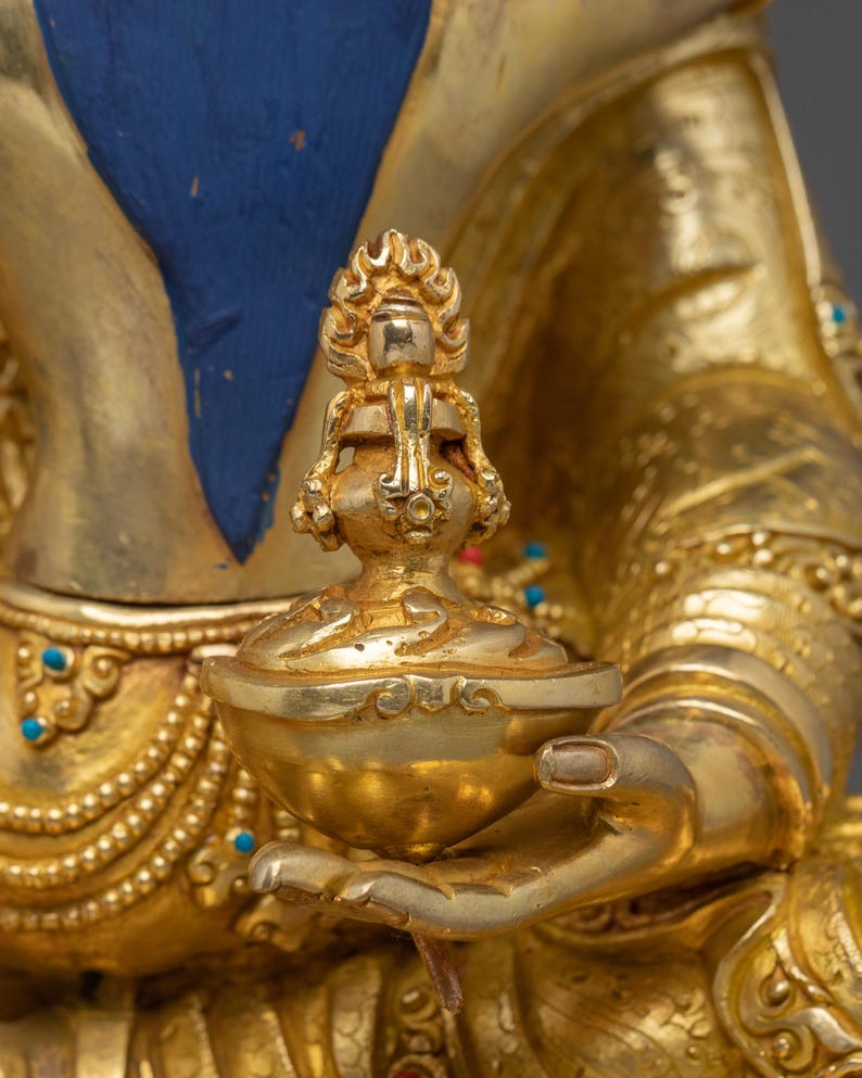 K&ouml;nnte beinhalten: Nahaufnahme einer goldenen religi&ouml;sen Statue, die eine kleine, verzierte Schale h&auml;lt. Die Statue weist komplizierte Details auf, darunter ein flammenartiges Design auf der Schale und t&uuml;rkisfarbene Akzente. Der Hintergrund ist ein weiches, verschwommenes Gold.
