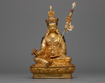 May include: A gold-colored statue of a deity sitting in a meditative pose. The statue is holding a scepter in its right hand and has a crown on its head. The statue is sitting on a lotus base.