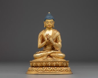 May include: A gold Buddha statue with a blue topknot. The seated figure has hands in a prayer position. The base is gold with a detailed floral design. The background is a solid grey colour. The statue is a representation of Buddhist art.
