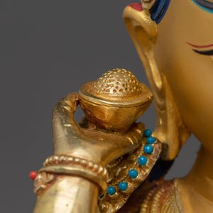 K&ouml;nnte beinhalten: Nahaufnahme einer goldfarbenen Statue, die eine Figur mit einer kleinen, verzierten Schale zeigt. Die Statue weist komplizierte Details auf, darunter ein Armband mit einer roten Perle und eine Halskette mit t&uuml;rkisfarbenen Perlen. Das Haar der Figur ist mit blauen Akzenten verziert.