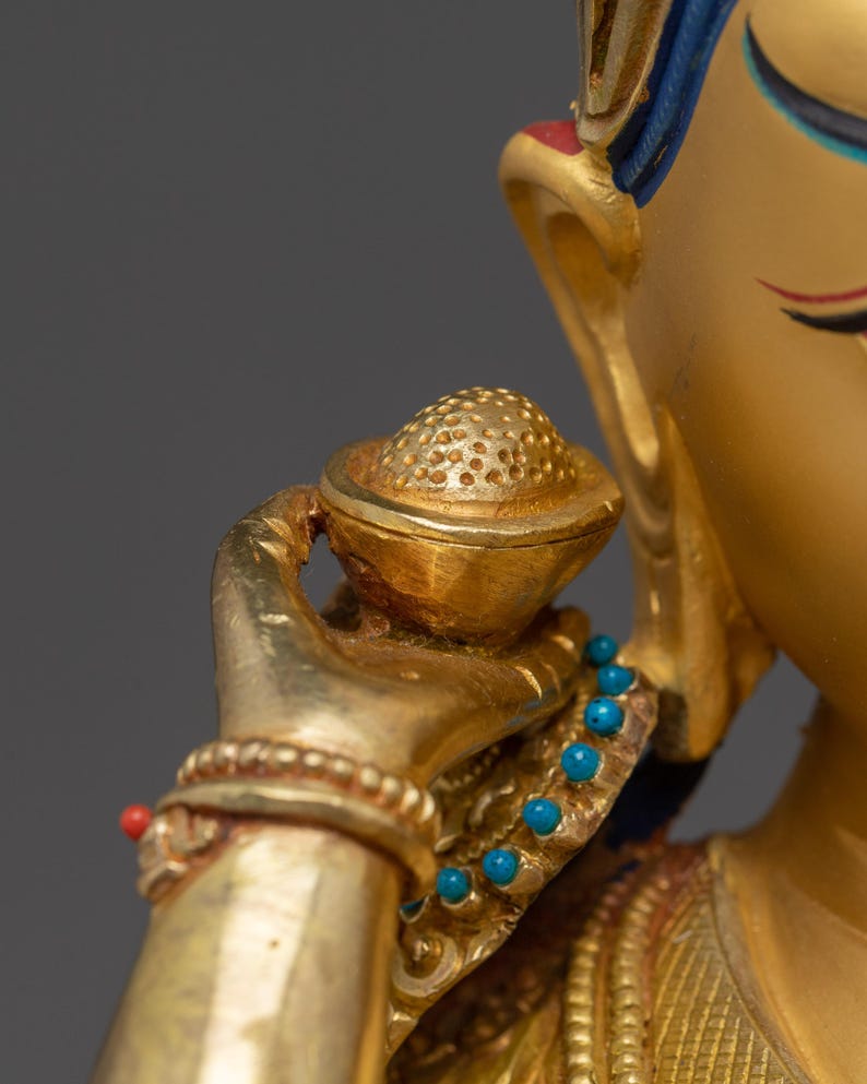 K&ouml;nnte beinhalten: Nahaufnahme einer goldfarbenen Statue, die eine Figur mit einer kleinen, verzierten Schale zeigt. Die Statue weist komplizierte Details auf, darunter ein Armband mit einer roten Perle und eine Halskette mit t&uuml;rkisfarbenen Perlen. Das Haar der Figur ist mit blauen Akzenten verziert.