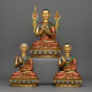 Puede incluir: Tres estatuas doradas de figuras budistas en posición de meditación sentada. Las estatuas están adornadas con intrincados detalles y pintura colorida. La figura central es más grande que las dos figuras laterales.