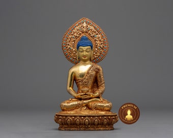 May include: A gold-coloured statue of Buddha sitting in meditation with a blue crown and a gold-coloured base. The statue is on a grey background.