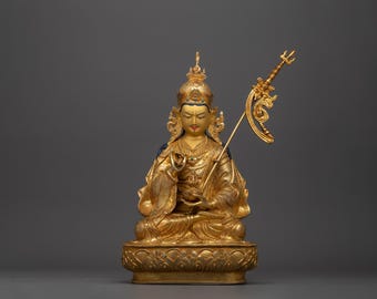 May include: A gold-colored statue of a seated figure with a crown and a trident. The figure is wearing a robe and holding a hand gesture. The statue is on a lotus base.