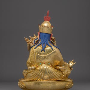 K&ouml;nnte beinhalten: Goldene Statue einer sitzenden Figur, von hinten gesehen. Die Figur hat einen blauen Kopf und flie&szlig;endes blaues Haar, das eine goldene Krone mit roten Akzenten tr&auml;gt. Das Gewand ist goldfarben mit aufwendigen Mustern, und der Sockel ist ebenfalls goldfarben.