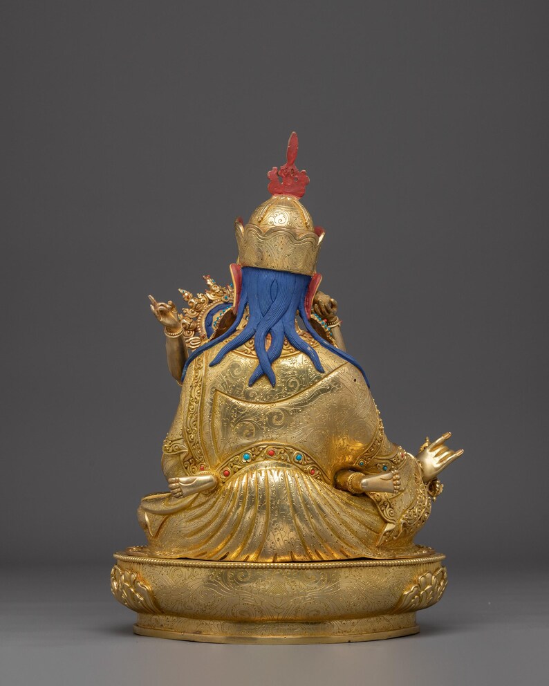 K&ouml;nnte beinhalten: Goldene Statue einer sitzenden Figur, von hinten gesehen. Die Figur hat einen blauen Kopf und flie&szlig;endes blaues Haar, das eine goldene Krone mit roten Akzenten tr&auml;gt. Das Gewand ist goldfarben mit aufwendigen Mustern, und der Sockel ist ebenfalls goldfarben.