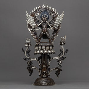 May include: A silver and bronze statue of a deity with wings, standing on a lotus base with a flame-like halo. The statue is adorned with intricate details and has a star-shaped ornament on the top.