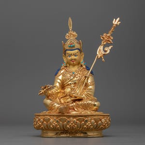 May include: A gold-colored statue of a seated figure with a crown and a scepter. The figure is adorned with turquoise and blue accents. The statue is sitting on a lotus-shaped base.
