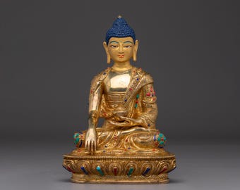 May include: A gold Buddha statue with a blue head and detailed craftsmanship. The seated figure has a calm expression and is decorated with colourful gemstones. The statue sits on a decorative base, displaying a mix of artistry and spirituality.