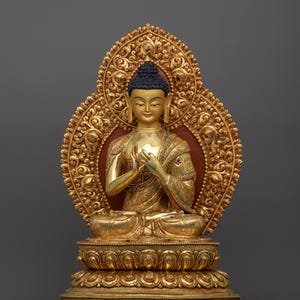 K&ouml;nnte beinhalten: Eine goldfarbene Buddha-Statue, die in meditativer Haltung sitzt. Die Statue ist mit aufwendigen Details verziert und vor einem grauen Hintergrund platziert.