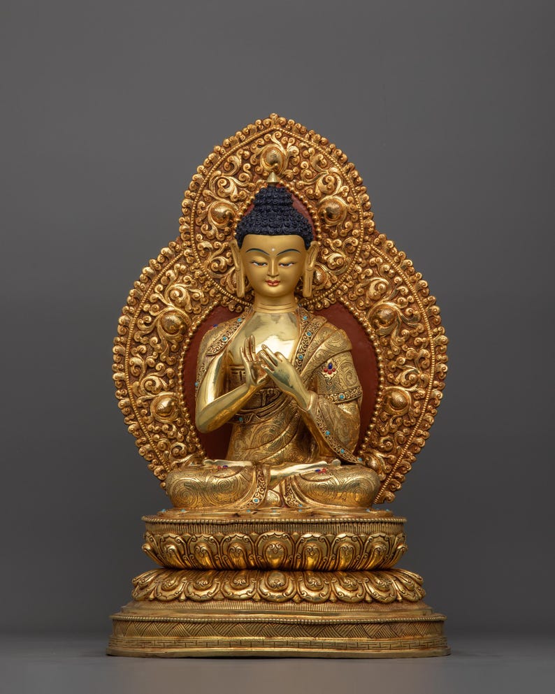 K&ouml;nnte beinhalten: Eine goldfarbene Buddha-Statue, die in meditativer Haltung sitzt. Die Statue ist mit aufwendigen Details verziert und vor einem grauen Hintergrund platziert.