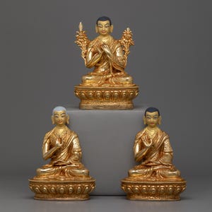 Puede incluir: Tres estatuas doradas de figuras budistas en posición de meditación sentada. Las estatuas están sobre una superficie gris.