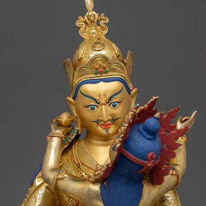 K&ouml;nnte beinhalten: Goldfarbene Statue einer Gottheit mit einem juwelenbesetzten Diadem und aufwendigen Details. Die Gottheit hat einen ruhigen Ausdruck, blaue Augen und einen roten Schnurrbart. Die Gottheit h&auml;lt ein blaues und rotes Objekt. Grauer Hintergrund.