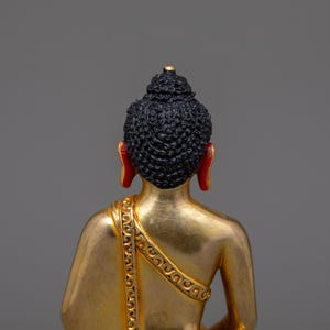 May include: A gold-colored Buddha statue with a black hair and red earlobes. The statue is sitting in a meditative pose.
