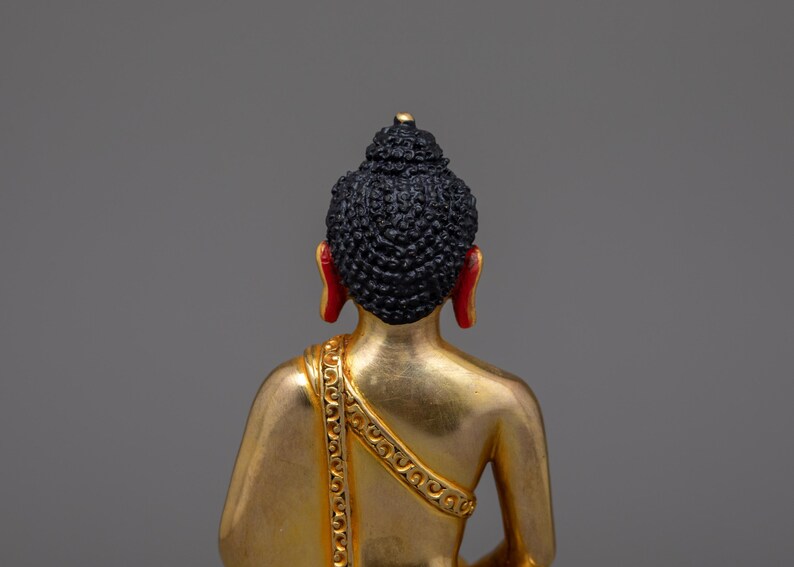 May include: A gold-colored Buddha statue with a black hair and red earlobes. The statue is sitting in a meditative pose.