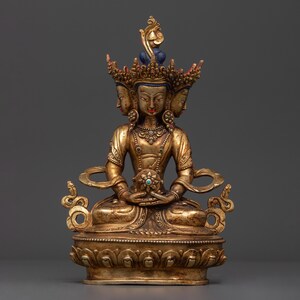 May include: A gold-coloured statue of a deity with three faces and a crown. The deity is seated in a meditative pose, holding a jeweled orb in their hands. The statue is decorated with intricate details and is set on a lotus base.