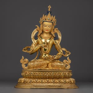 Könnte beinhalten: Eine goldene Statue einer sitzenden Gottheit mit aufwendigen Details. Die Figur trägt eine Krone und Schmuck, mit einem friedlichen Ausdruck. Die Statue steht auf einem verzierten Sockel, der eine Mischung aus Kunst und Spiritualität zeigt. Das Design ist reich an Details und Farben.