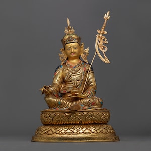 May include: A gold-colored statue of a deity sitting in a meditative pose. The statue is adorned with intricate details and colorful gemstones. The deity is holding a staff in their right hand and has a serene expression on their face.