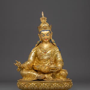 K&ouml;nnte beinhalten: Eine goldene Statue einer sitzenden Figur, geschm&uuml;ckt mit einer detaillierten Krone und Gew&auml;ndern. Die Figur befindet sich in einer meditativen Pose und h&auml;lt Gegenst&auml;nde in ihren H&auml;nden. Die Statue steht auf einem dekorativen Sockel mit aufwendigen Schnitzereien und Details.