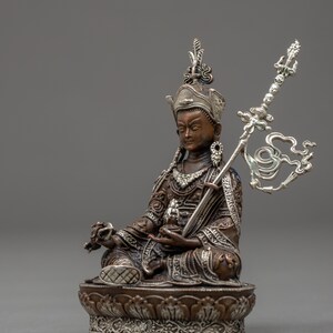 May include: A silver and bronze statue of a deity sitting in a meditative pose. The deity is wearing a crown and ornate clothing. The statue is holding a scepter in one hand and a lotus flower in the other.
