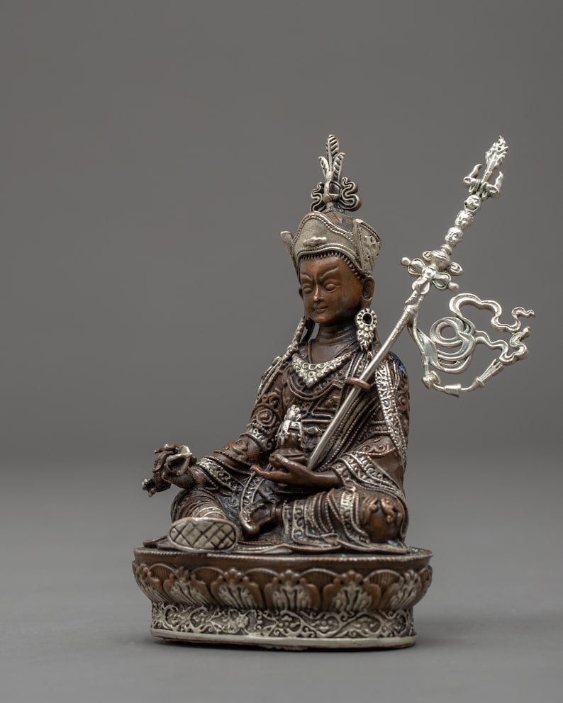 May include: A silver and bronze statue of a deity sitting in a meditative pose. The deity is wearing a crown and ornate clothing. The statue is holding a scepter in one hand and a lotus flower in the other.