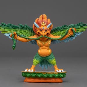 Puede incluir: Una estatua colorida de una criatura mítica con alas, un pico de pájaro y una serpiente en sus garras. La criatura es naranja, verde y azul, y está de pie sobre una base verde y naranja.