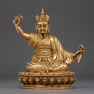 May include: A gold-colored statue of a deity sitting in a meditative pose. The statue is adorned with intricate details and is holding a scepter in one hand and a ritual dagger in the other. The deity is wearing a crown and ornate clothing.
