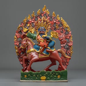 Könnte beinhalten: Eine handbemalte Statue einer blauhäutigen Gottheit auf einem braunen Pferd vor einer rot-goldenen, flammenförmigen Mandorla. Die Figur trägt eine goldene Krone und farbenfrohe Gewänder und steht auf einem grünen Sockel mit Bergreliefs.
