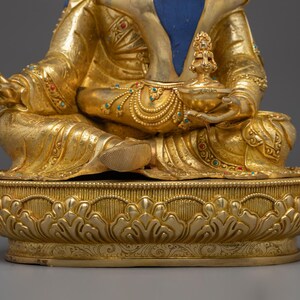 K&ouml;nnte beinhalten: Eine goldene Buddha-Statue in sitzender Position, verziert mit aufwendigen Details. Die Statue hat einen blauen Bereich auf der Brust und ist mit farbigen Edelsteinen verziert. Der Sockel ist aufwendig mit einem floralen Design geschnitzt, das Kunstfertigkeit und Spiritualit&auml;t zeigt.