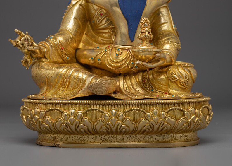 K&ouml;nnte beinhalten: Eine goldene Buddha-Statue in sitzender Position, verziert mit aufwendigen Details. Die Statue hat einen blauen Bereich auf der Brust und ist mit farbigen Edelsteinen verziert. Der Sockel ist aufwendig mit einem floralen Design geschnitzt, das Kunstfertigkeit und Spiritualit&auml;t zeigt.