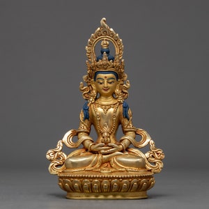 Può includere: Una statua dorata di un Buddha seduto con una corona blu e un fiore di loto nelle mani. La statua è su una base con dettagli intricati.